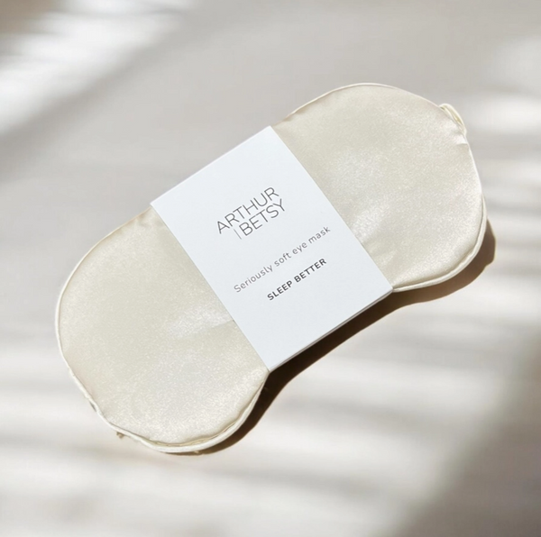 An Arthur Betsy sleep mask made of soft, lustrous champagne-colored fabric, wrapped in a white paper band with the brand name 'ARTHUR BETSY' printed in black. Part of the Sleepover Macaron Gift Set.