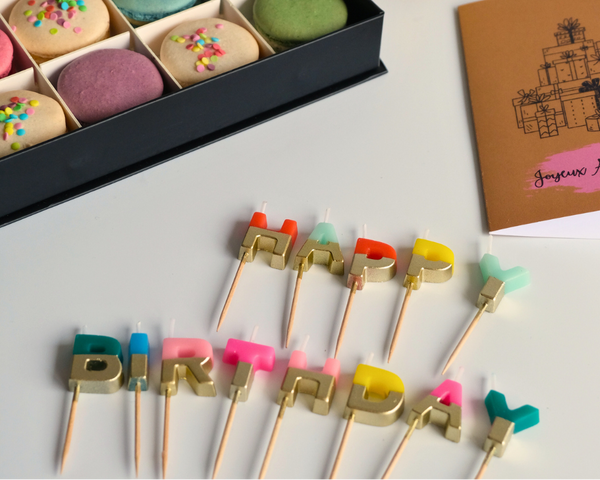 Colorful birthday-themed cookies in a box with 'Happy Birthday' candles on a white surface.
