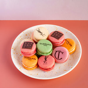 Assorted macarons on a white plate with visible brand logos against a pink background