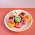 Assorted macarons on a white plate with visible brand logos against a pink background