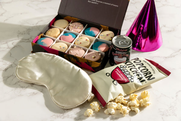 A curated gift set featuring a box of twelve assorted macarons, a can of Clover Club cocktail, a cream satin sleep mask, and gourmet popcorn. The perfect addition to a sleepover night.
