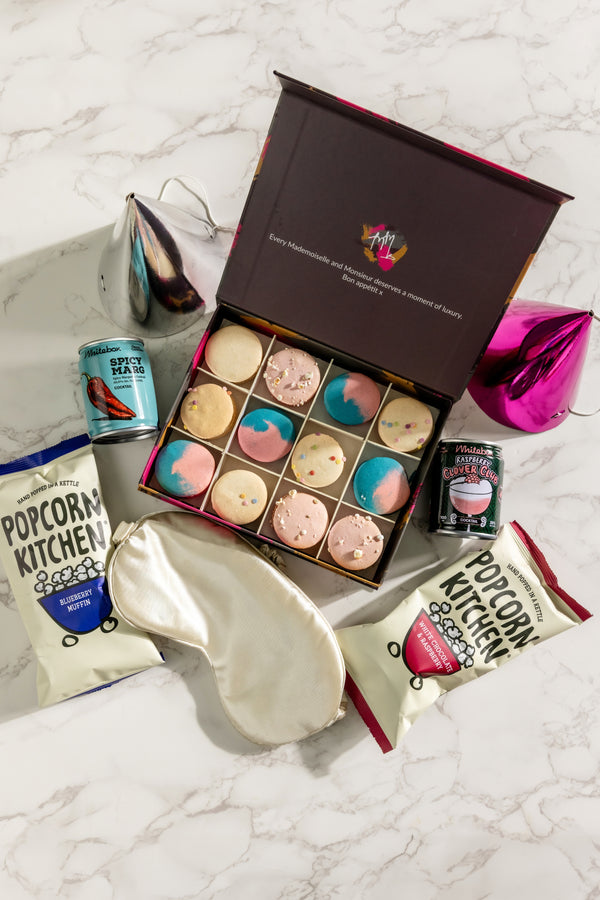 Gourmet gift set on a white marble surface with colourful macarons in an open box, a satin sleep mask, gourmet popcorn and two cans of cocktails. The perfect celebration gift set! 
