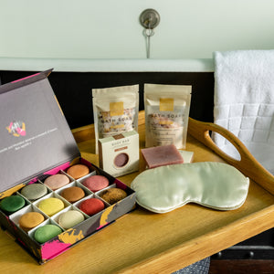 A bath and beauty gift set arranged on a wooden tray beside a bathtub. The set includes an open box of assorted colorful bath macarons, two pouches of Bath Soap, a small boxed item, a pale green satin eye mask, and a folded white towel hanging on the tub. 