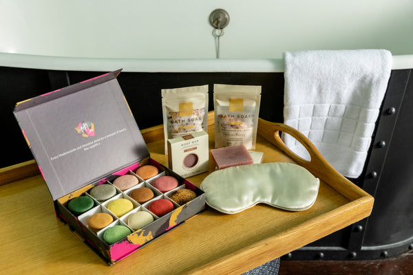 A bath and beauty gift set arranged on a wooden tray beside a bathtub. The set includes an open box of assorted colorful bath macarons, two pouches of Bath Soap, a small boxed item, a pale green satin eye mask, and a folded white towel hanging on the tub. 