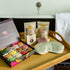 A bath and beauty gift set arranged on a wooden tray beside a bathtub. The set includes an open box of assorted colorful bath macarons, two pouches of Bath Soap, a small boxed item, a pale green satin eye mask, and a folded white towel hanging on the tub. 
